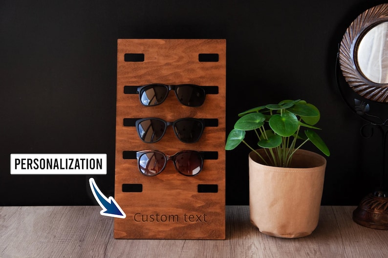 Puede incluir: Un soporte de madera marr&oacute;n oscuro para gafas de sol con tres pares de gafas de sol. El soporte tiene ranuras para sujetar las gafas. El texto "Personalizaci&oacute;n" y "Custom text" son visibles.
