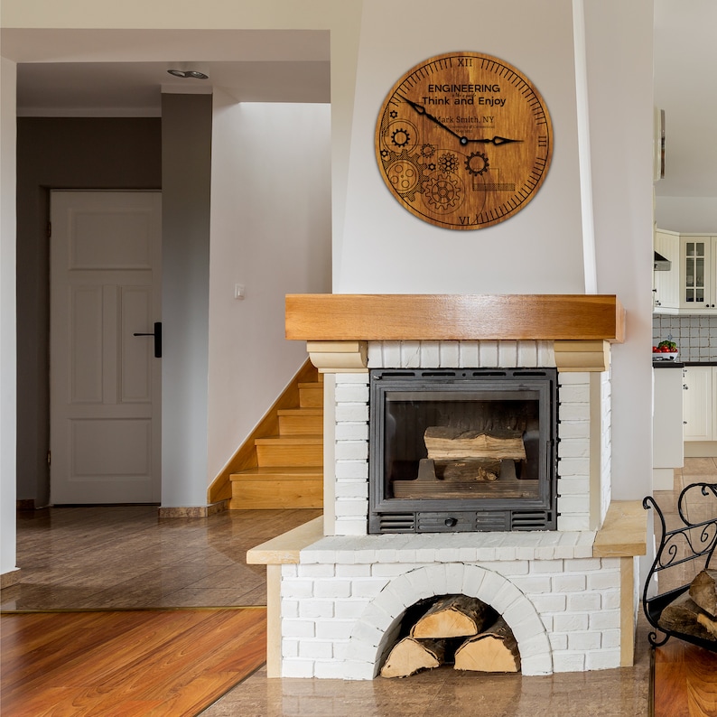May include: A wooden clock with a brown background and black gears and hands. The clock face reads "Engineering Think and Enjoy" with the text "Mark Smith, NY" below. The clock is hanging on a white wall above a white brick fireplace.