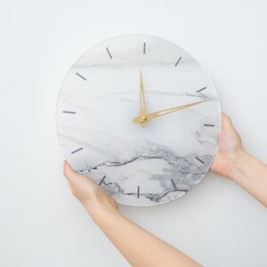 May include: A white marble clock with gold hands. The clock is being held by two hands.