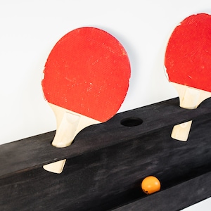 May include: Two red ping pong paddles with black handles are mounted on a black wooden rack. An orange ping pong ball is in the rack.