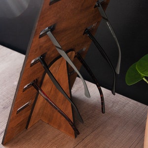 Puede incluir: Un expositor de gafas de madera con varios pares de gafas. El expositor es de madera marr&oacute;n oscuro y tiene ranuras para sujetar las gafas. Las gafas tienen varios estilos y colores de montura, incluyendo negro, gris y carey.