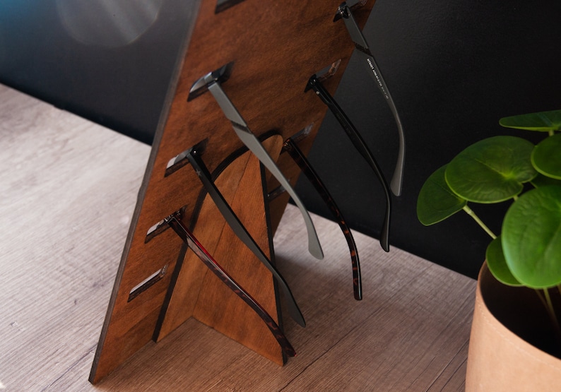 Puede incluir: Un expositor de gafas de madera con varios pares de gafas. El expositor es de madera marr&oacute;n oscuro y tiene ranuras para sujetar las gafas. Las gafas tienen varios estilos y colores de montura, incluyendo negro, gris y carey.