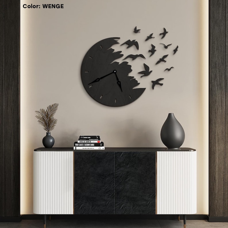 May include: A black wall clock with a unique design featuring a circular clock face with a distressed edge and birds in flight. The clock is mounted on a neutral-colored wall. The text "Color: WENGE" is in the top left corner.