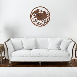 May include: A brown wooden wall clock with a silhouette of two birds perched on a branch with leaves. The clock is hanging on a white wall above a white sofa.