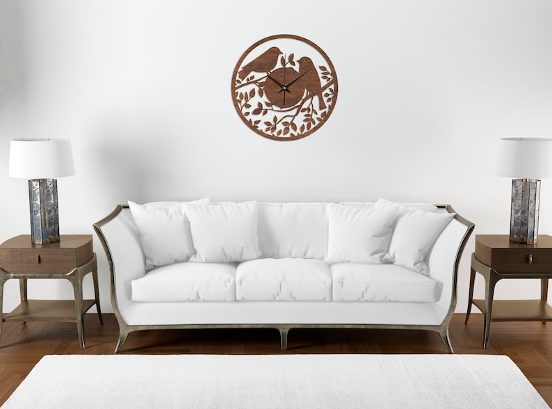 May include: A brown wooden wall clock with a silhouette of two birds perched on a branch with leaves. The clock is hanging on a white wall above a white sofa.