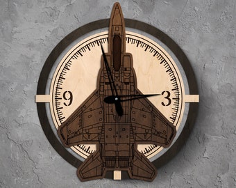 Könnte beinhalten: Eine Holzuhr mit braunem Hintergrund und schwarzem Rahmen. Das Zifferblatt der Uhr ist aus hellem Holz und zeigt eine schwarze Silhouette eines Kampfjets. Die Zahlen 9 und 3 sind auf dem Zifferblatt sichtbar.