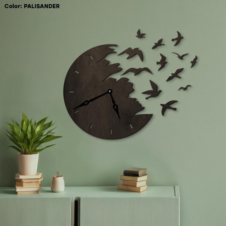 May include: A dark brown wooden wall clock with a unique design. The clock face is partially broken, with birds flying out from the broken section. The clock has black hands and is mounted on a green wall. The color is Palissander.