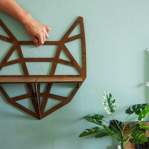 May include: A wooden geometric fox-shaped shelf with a brown stain. The shelf is mounted on a light blue wall.