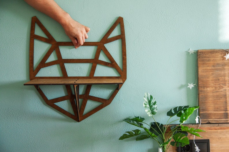 May include: A wooden geometric fox-shaped shelf with a brown stain. The shelf is mounted on a light blue wall.