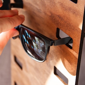 Puede incluir: Gafas de sol negras con montura rectangular se colocan en un expositor de madera. El expositor tiene ranuras para cada par de gafas. Las gafas de sol se est&aacute;n colocando en el expositor.