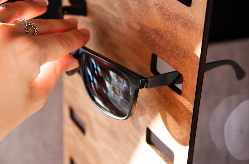 Puede incluir: Gafas de sol negras con montura rectangular se colocan en un expositor de madera. El expositor tiene ranuras para cada par de gafas. Las gafas de sol se est&aacute;n colocando en el expositor.