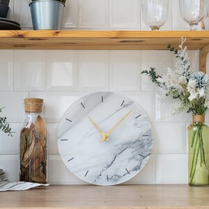 May include: A white marble clock with gold hands and black markings. The clock is round and has a white background with gray marble veins.