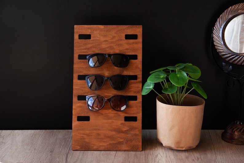 Puede incluir: Un organizador de gafas de sol de madera con tres pares de gafas de sol. El organizador est&aacute; te&ntilde;ido de un marr&oacute;n c&aacute;lido y tiene barras negras para sujetar las gafas. A la derecha hay una peque&ntilde;a planta en maceta en una maceta de papel.