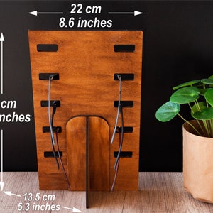 Puede incluir: Un expositor de gafas de madera, de 38 cm de alto y 22 cm de ancho, con acabado de madera natural. El expositor tiene ranuras para sujetar las gafas y una base de soporte. A la derecha hay una planta en maceta.