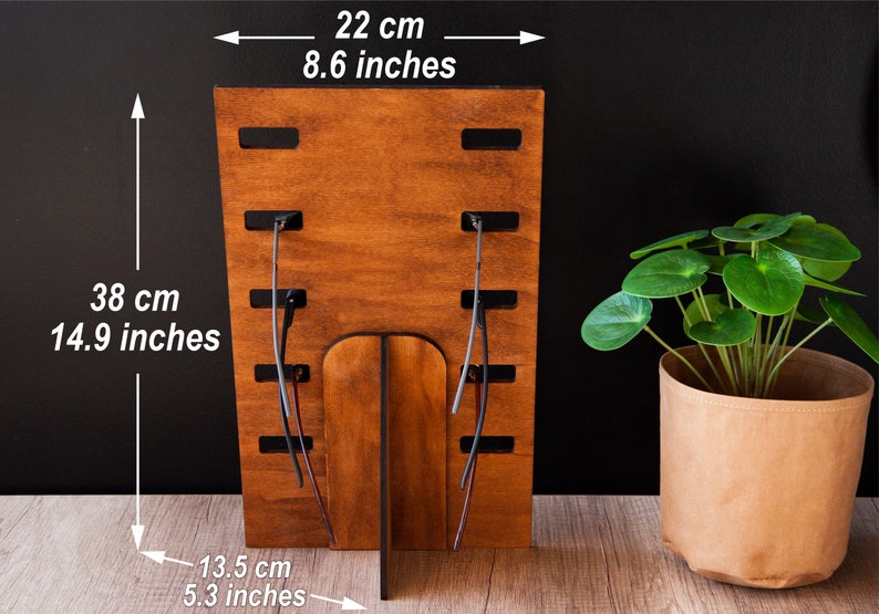 Puede incluir: Un expositor de gafas de madera, de 38 cm de alto y 22 cm de ancho, con acabado de madera natural. El expositor tiene ranuras para sujetar las gafas y una base de soporte. A la derecha hay una planta en maceta.