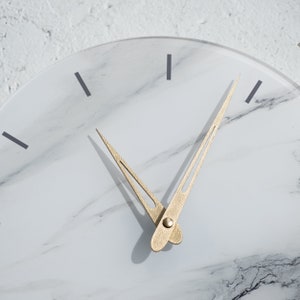 May include: A white marble clock face with gold-colored hour and minute hands. The clock face has a subtle marbled pattern.