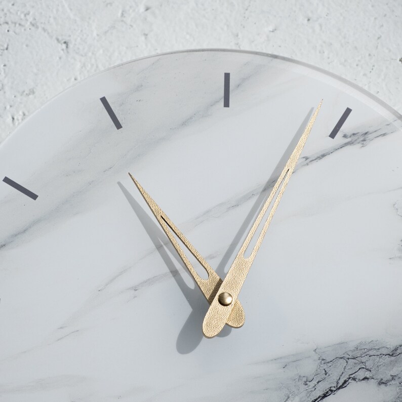 May include: A white marble clock face with gold-colored hour and minute hands. The clock face has a subtle marbled pattern.