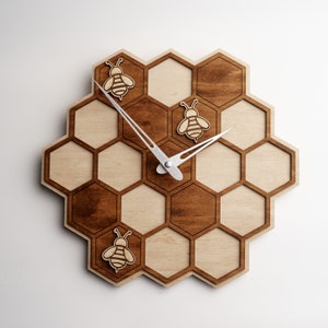 Puede incluir: Un reloj de madera en forma de panal con agujas plateadas y tres diseños de abejas. El reloj está hecho de madera clara y oscura y tiene un aspecto rústico.