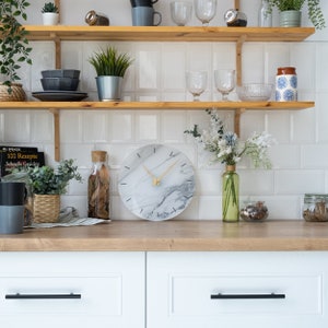 May include: A white marble clock with black markings and gold hands hangs on a wooden shelf in a kitchen. The clock is surrounded by plants, books, and other decorative items.