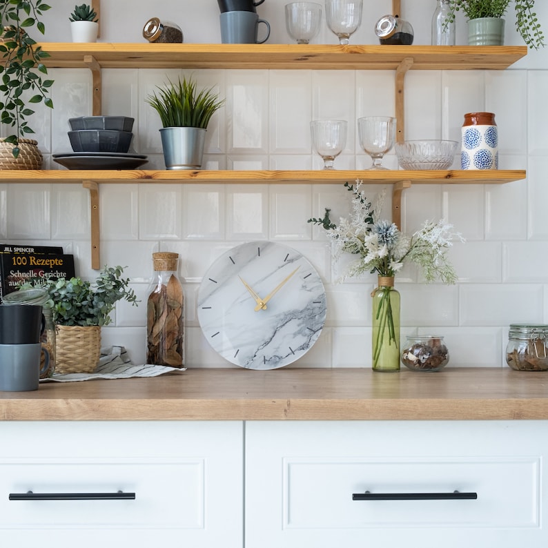 May include: A white marble clock with black markings and gold hands hangs on a wooden shelf in a kitchen. The clock is surrounded by plants, books, and other decorative items.