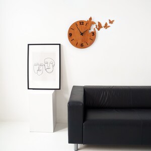 May include: A wooden clock with a butterfly design. The clock is brown and has black numbers. The clock is hanging on a white wall. There is a black leather sofa in the background.