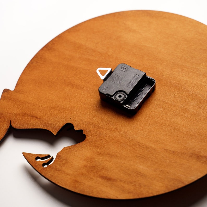 May include: The back of a wooden clock with a black clock mechanism. The clock is shaped like a crescent moon with a silhouette of a bird flying out of the moon.