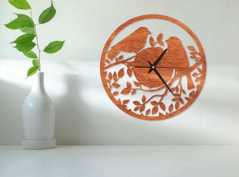 May include: A wooden wall clock with a brown wood finish. The clock face is surrounded by a design of two birds perched on a branch with leaves. The clock hands are black.