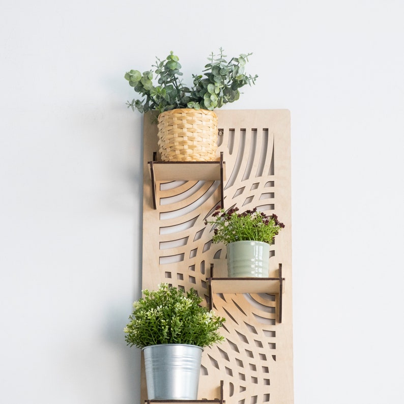 May include: A wooden wall shelf with a geometric design features three potted plants. The top shelf has a green plant in a woven basket, the middle shelf has a green plant in a light green pot, and the bottom shelf has a green plant in a silver pot.