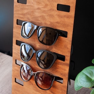 Puede incluir: Un expositor de gafas de sol de madera con tres pares de gafas de sol. El expositor es de madera marr&oacute;n claro y tiene soportes negros para las gafas. Las gafas de sol tienen monturas negras y lentes tintadas.
