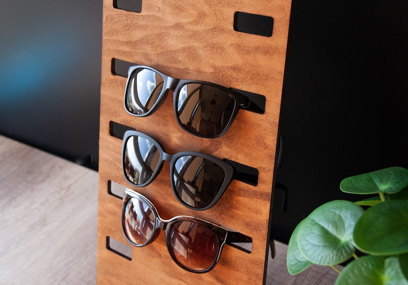 Puede incluir: Un expositor de gafas de sol de madera con tres pares de gafas de sol. El expositor es de madera marr&oacute;n claro y tiene soportes negros para las gafas. Las gafas de sol tienen monturas negras y lentes tintadas.