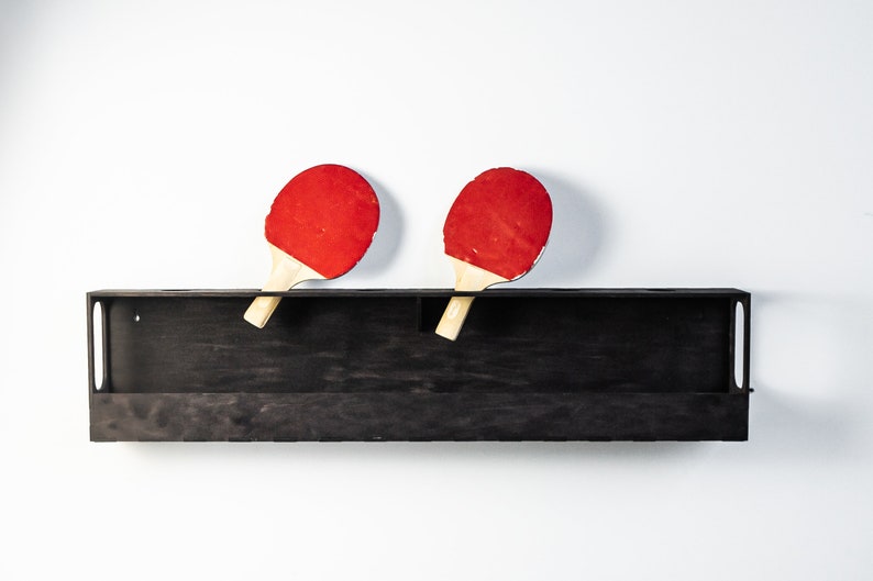 May include: Two red and black ping pong paddles are hanging on a black wooden shelf against a white wall.