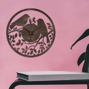 May include: A brown wooden clock with a silhouette of two birds perched on a branch with leaves. The clock has a simple design with black hands and a white face.