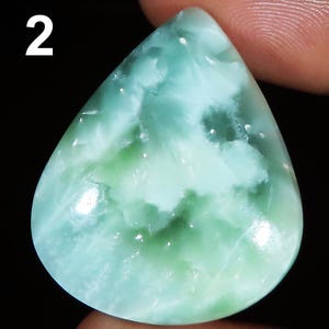 Può includere: Una gemma a forma di goccia con una miscela di tonalità azzurre e verdi. La pietra ha un aspetto nuvoloso e marmorizzato con aree di tonalità più chiare e più scure. Il numero "2" è visibile nell'angolo in alto a sinistra.