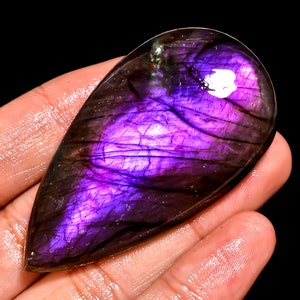 Puede incluir: Una gema pulida en forma de lágrima, con un vibrante tono púrpura. La piedra muestra juegos de luz internos, con vetas de púrpura más claro y un borde oscuro, casi negro. La gema se sostiene en una mano sobre un fondo negro.