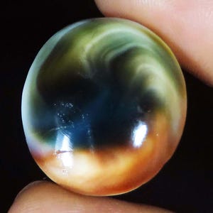 May include: A round, polished gemstone cabochon featuring a smooth, swirling pattern of deep forest green, dark navy blue, and warm amber-orange tones. The surface has a high-gloss finish that reflects light, creating bright white highlights.