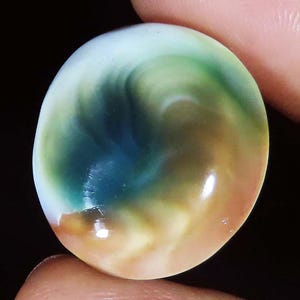 May include: A round, polished cabochon gemstone featuring a smooth, swirling pattern of deep forest green, dark teal, and warm amber tones. The surface has a high-gloss finish that reflects light, creating a luminous, multi-coloured appearance.