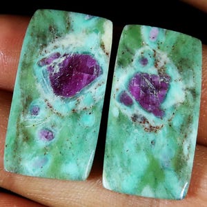 Peut inclure: Deux pierres précieuses rectangulaires. Elles présentent une base turquoise avec des marbrures vertes et marron. Chaque pierre a une inclusion centrale, facettée, magenta foncé.