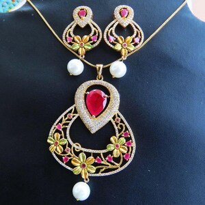 May include: A gold-toned jewelry set featuring a pendant necklace and matching earrings. The pendant has a large, teardrop-shaped red gemstone surrounded by smaller clear stones. The earrings and pendant are adorned with floral and pearl accents.
