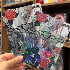 May include: Two rectangular holographic prints featuring a graphic illustration of two skeletons embracing, red roses, a heart-shaped chain, and a blindfolded figure. The surface has a reflective, shimmering finish that catches light with rainbow-colored sparkles.