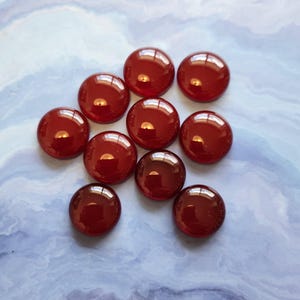 Peut inclure: Onze cabochons ronds en verre rouge brillant sont disposés sur une surface marbrée bleu clair et blanche. Les cabochons reflètent la lumière, leur donnant un aspect brillant. Ces cabochons décoratifs en verre sont parfaits pour l'artisanat et la décoration intérieure. Chaque cabochon mesure environ 2 cm de diamètre.