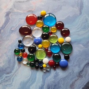 May include: A collection of colourful glass marbles in various shades of red, blue, green, yellow, and clear. The marbles are arranged in a loose, organic pattern on a light blue and white marbled surface.