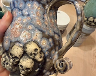 May include: A handmade ceramic mug with a unique design. The mug features a textured surface with a blue, brown, and gold glaze. Several small skull sculptures are attached to the base of the mug. The handle has a decorative swirl.