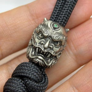 May include: A silver-toned metal bead shaped like a fierce, horned creature with a small green stone set in the forehead. The bead is threaded onto a thick, black braided cord that features a large, intricate knot at the bottom end.