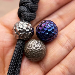 Op de afbeelding: Drie bolvormige kralen met een gehamerde, kuiltjesachtige textuur. Eén kraal is zilverkleurig, één is donker metallic grijs en één heeft een iriserende blauwe en paarse afwerking. Een zwart gevlochten koord is door de zilverkleurige kraal geknoopt.