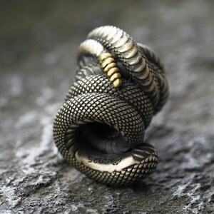 May include: A silver-toned ring in the shape of a coiled snake. The ring has a textured, scale-like pattern with a gold-colored accent. The ring is marked with "S925".