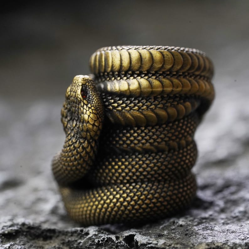 May include: A detailed, golden-colored snake sculpture with a coiled body and a detailed head. The snake's eye is a small red gem. The sculpture has a textured, scale-like pattern. The snake is coiled around an object, possibly a cup or a container.