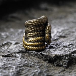 May include: A detailed, golden-colored ring shaped like a coiled snake. The ring features textured scales and a snake head, set against a textured, gray stone surface. The snake's body is wrapped around itself, creating a unique and eye-catching design.