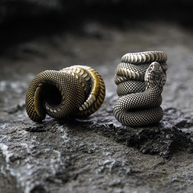 May include: Two metal snake rings. One ring is coiled in a circular shape, and the other is coiled vertically with a snake head at the top. The rings have a textured, scale-like pattern and are set on a gray stone surface.