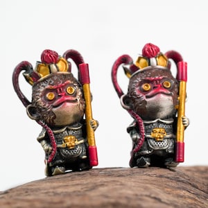 Pode incluir: Duas pequenas estatuetas detalhadas de macacos usando coroas vermelhas e douradas e segurando bastões. Os macacos têm detalhes vermelhos e dourados e são feitos de um metal escuro.