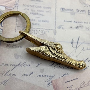 May include: A gold-colored alligator keychain. The alligator's head is detailed with textured scales and open jaws, attached to a round key ring. The keychain is on a vintage paper background.
